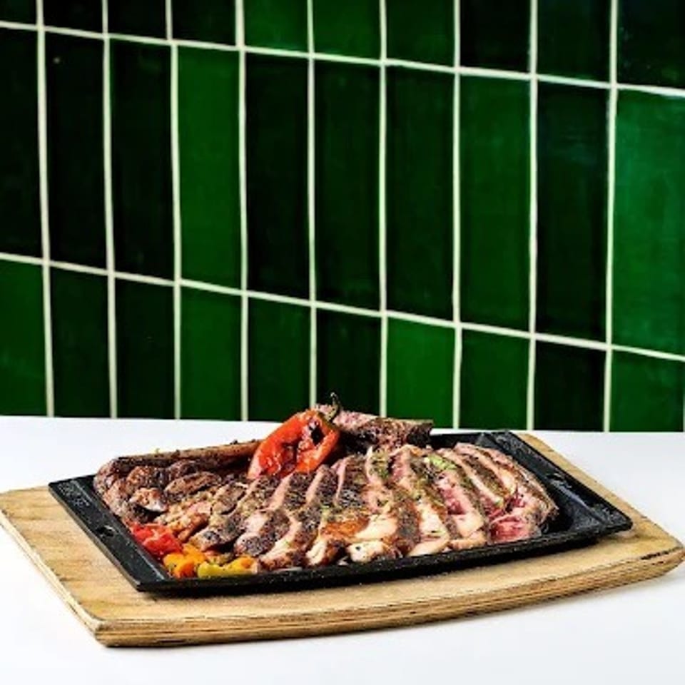 Grilled assorted meats and vegetables served on a black platter atop a wooden board against green tiled walls.