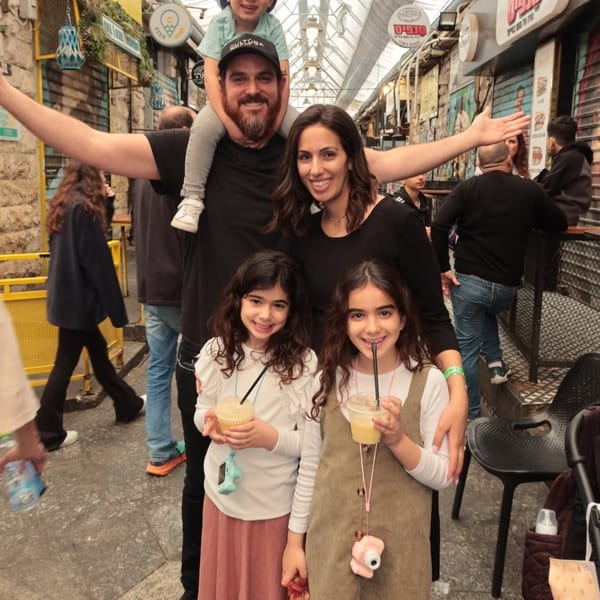 A family of five, including a father with a child on his shoulders, smiling in a bustling market.