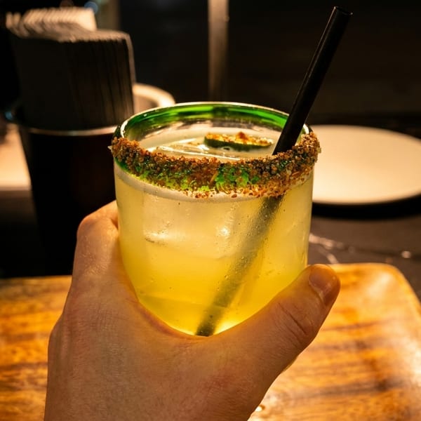 Hand holding a vibrant yellow cocktail with a spiced rim and lime slice in a dark restaurant.