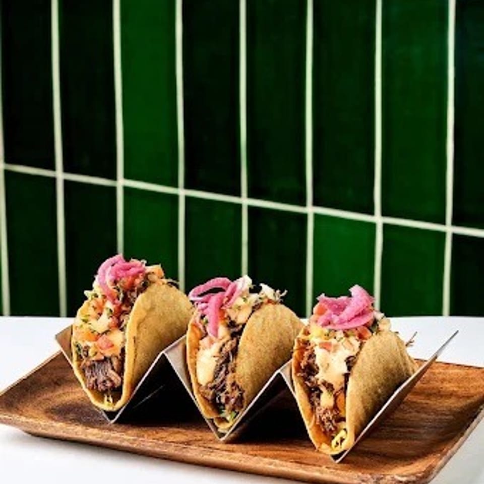 Three crispy tacos filled with meat, vegetables, and pink pickled onions on a wooden serving board against a dark green tiled wall.