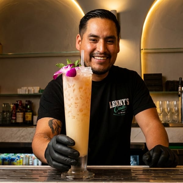 Smiling bartender in a black shirt and gloves presents a tall, garnished cocktail at a bar.