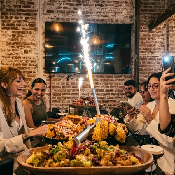 Friends gather around a table with large food platters and sparklers, smiling and taking photos.
