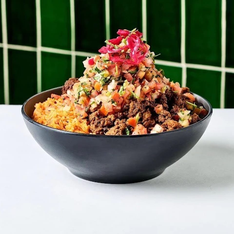 Loaded bowl of seasoned rice, ground meat, chopped vegetables, herbs, and pink pickled onions on a white table.