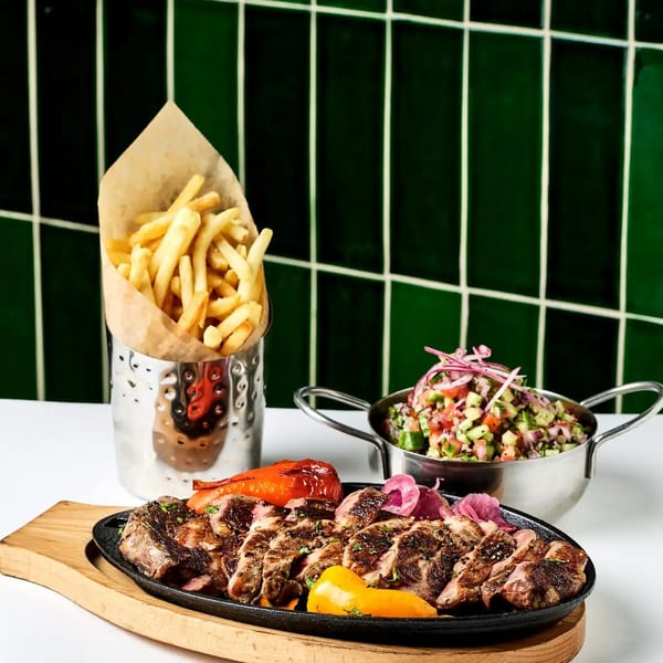 Sliced steak with roasted vegetables, fries in a metal cup, and a side salad on a white table.