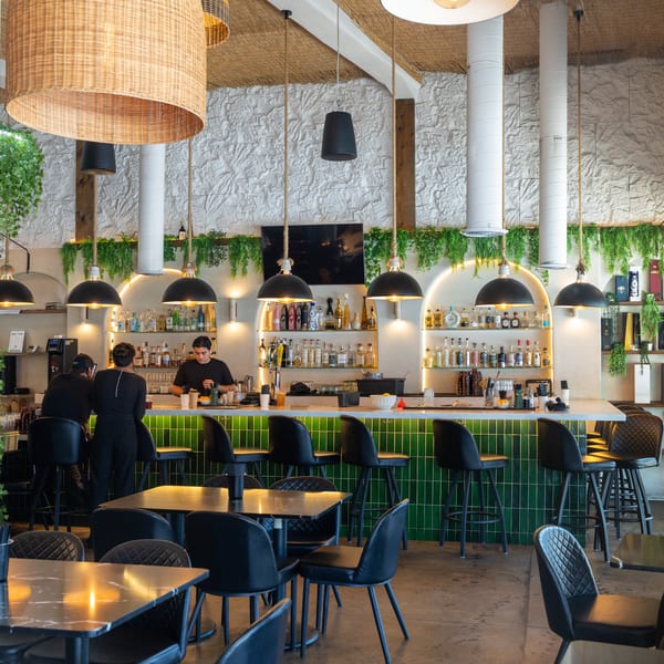 A vibrant restaurant interior with a green tiled bar, bottles, hanging plants, and black tables.