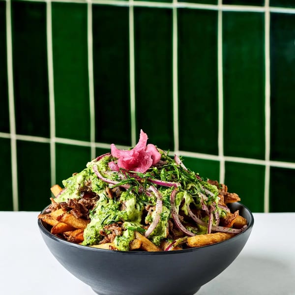 A black bowl filled with loaded fries, topped with green sauce, red onions, and pink pickled garnish.