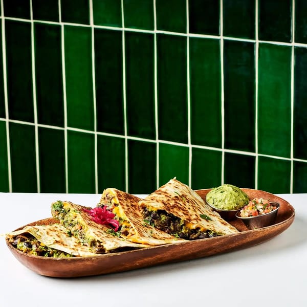 Quesadillas with guacamole and salsa on a wooden platter against a green tiled wall.