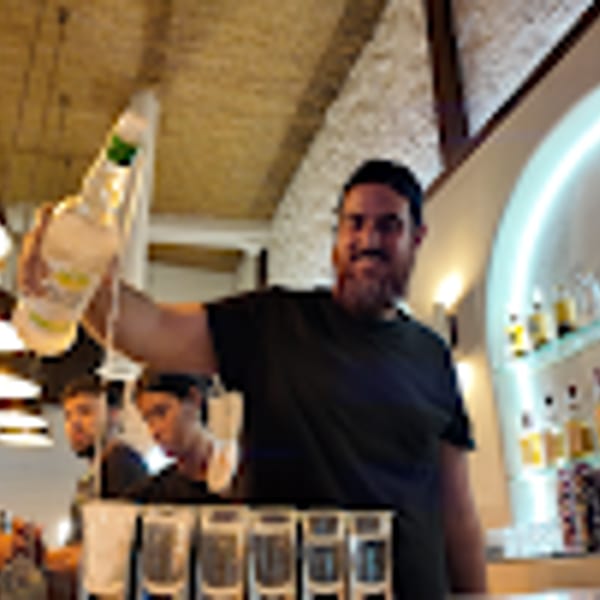 Smiling bartender pouring drinks into multiple shot glasses at a well-lit bar.