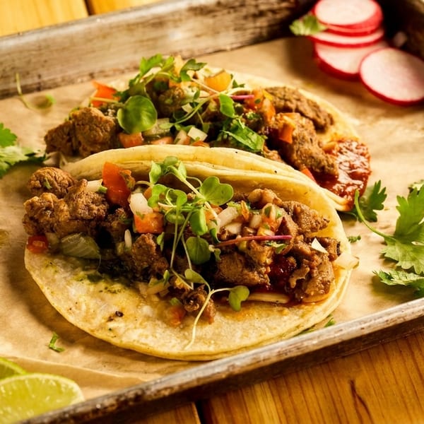Two tacos with meat, pico de gallo, and microgreens on a tray with limes and radishes.