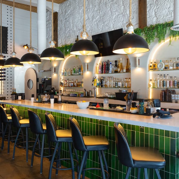 Modern bar with green tiled front, marble counter, black stools, and liquor bottles on arched shelves.