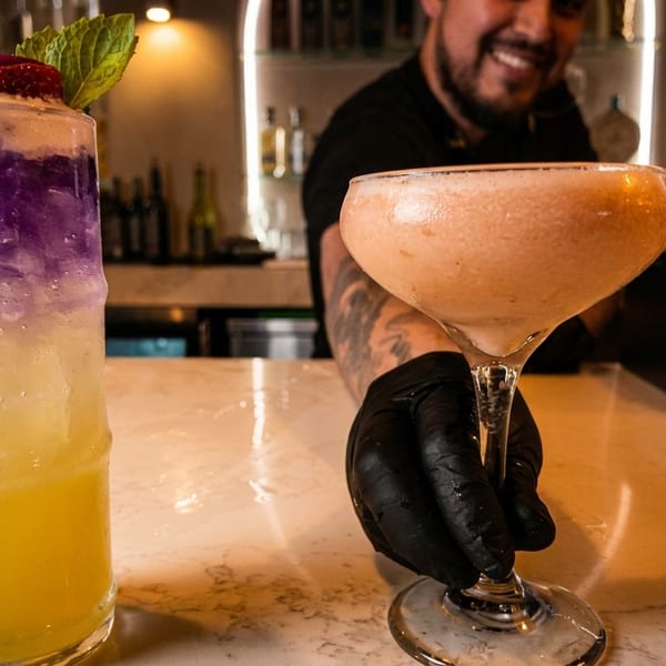 Bartender in black gloves holding a cocktail, with another colorful drink on the bar.