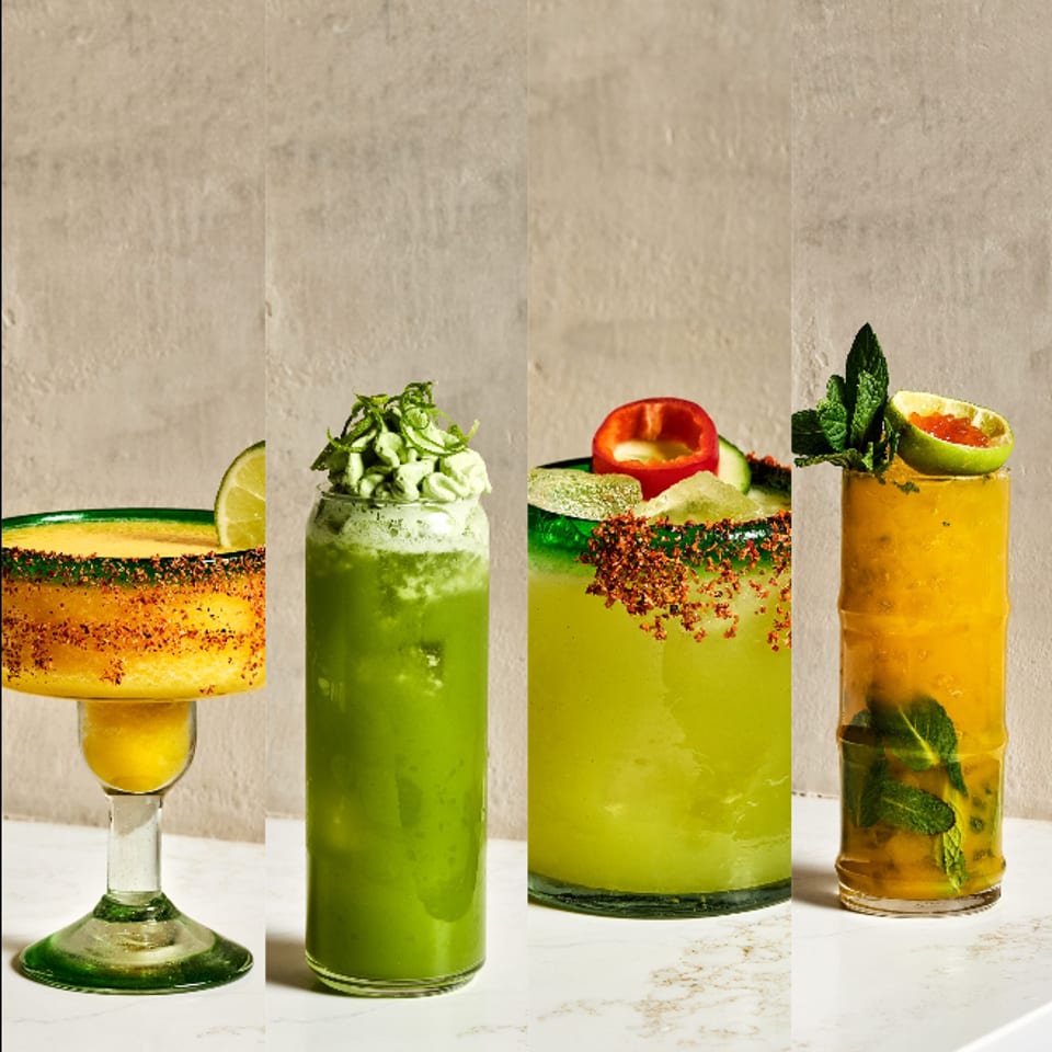 Four colorful cocktails with citrus garnishes, chili-salt rims, fresh herbs, and mint arranged on a marble table.