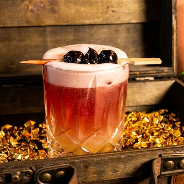 A cocktail with foam and cherries in a glass, presented in a wooden treasure chest filled with gold.