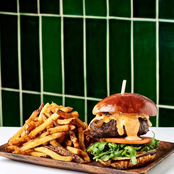 A juicy burger with sauce, tomato, and arugula, next to a pile of golden fries on a wooden tray.