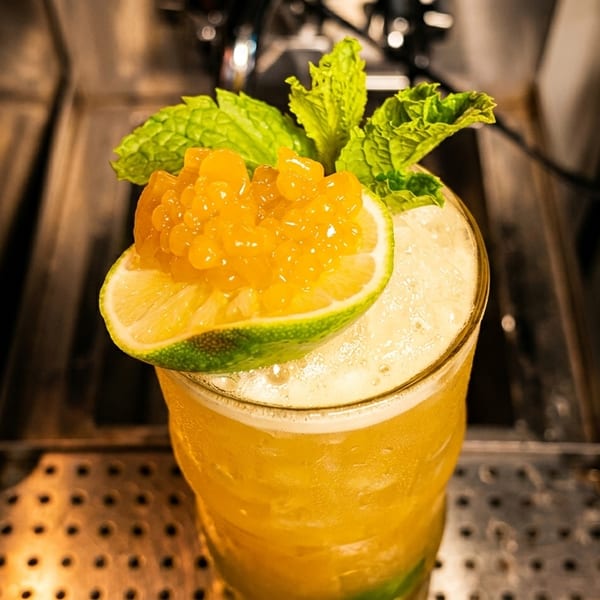 A vibrant yellow cocktail garnished with a lime wedge, passion fruit pulp, and fresh mint leaves.