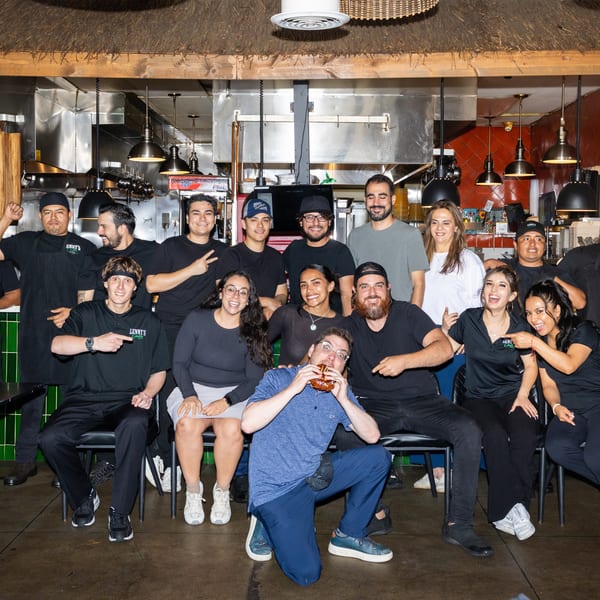 A large group of restaurant staff and a customer eating a burger pose in a kitchen.